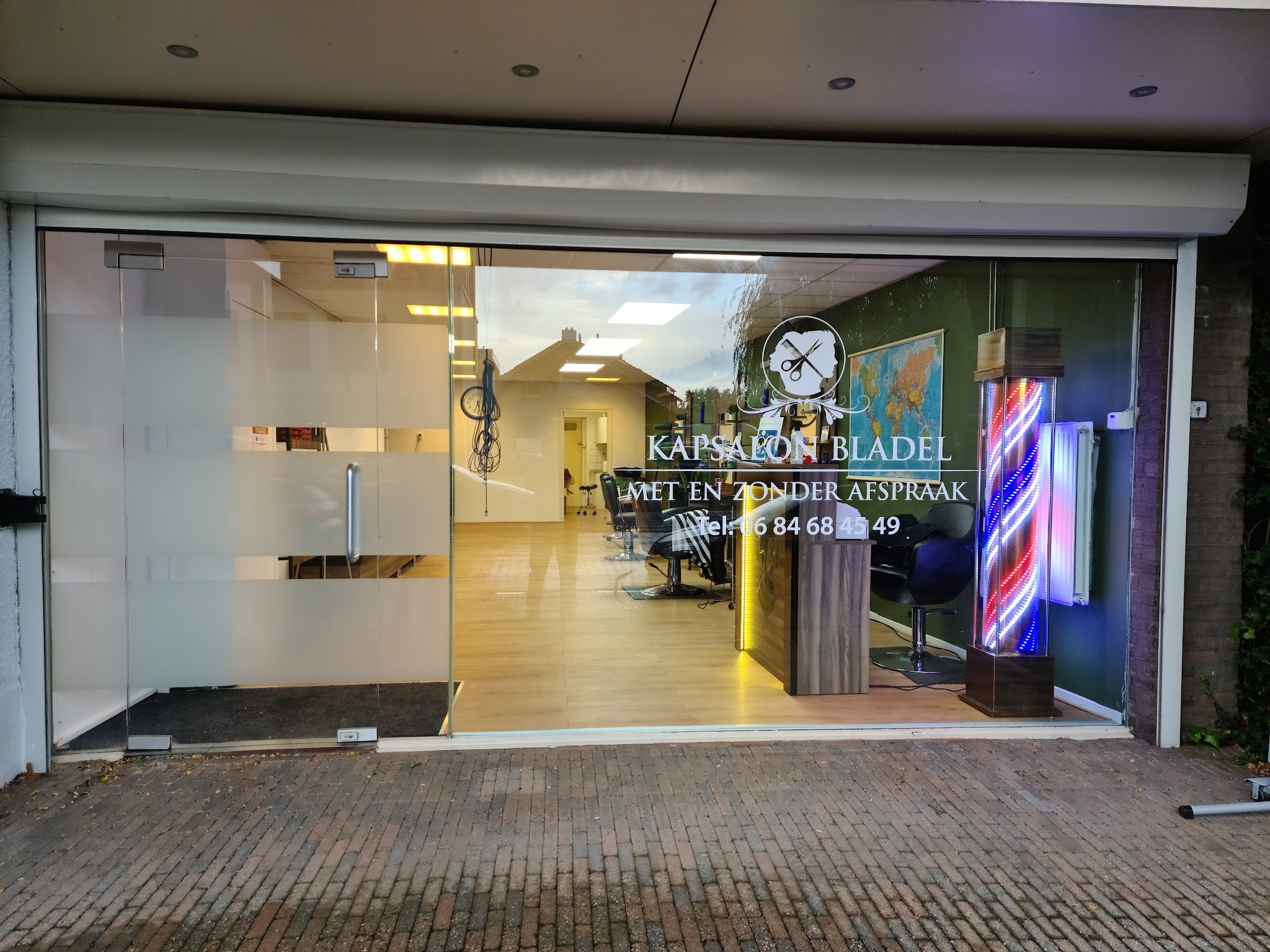 De gevel van Kapsalon Bladel aan de Sniederslaan in de avond, met de barberpaal en het raam met het logo